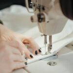 Detailed view of a sewing machine stitching fabric with visible hands, nails painted black.
